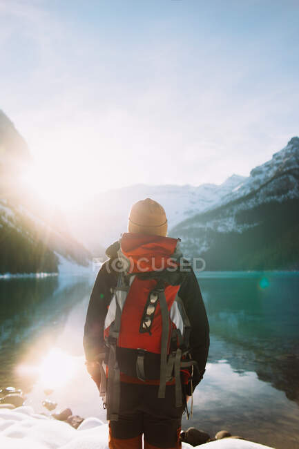 Vista posterior de caminante anónimo con mochila caminando contra el tranquilo lago Louise y las montañas nevadas en la soleada mañana de invierno en el Parque Nacional Banff - foto de stock