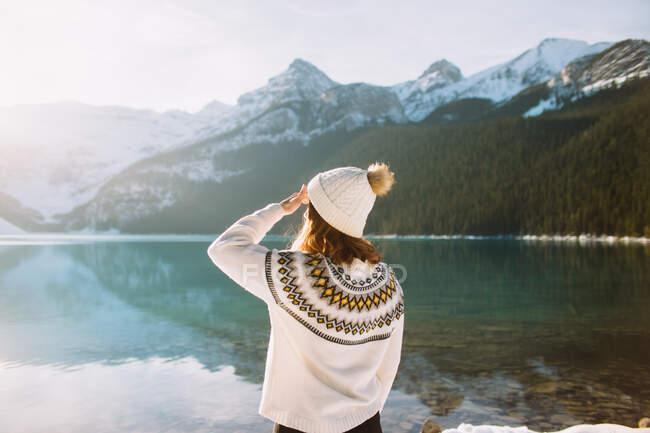 Rückansicht einer anonymen Wanderin in warmer Kleidung, die an einem sonnigen Wintermorgen im Banff National Park gegen den ruhigen Lake Louise und die stehenden Berge spaziert — Stockfoto