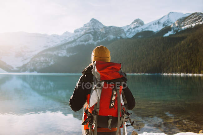 Vue arrière d'un randonneur anonyme avec sac à dos marchant contre le calme du lac Louise et les montagnes enneigées le matin ensoleillé d'hiver dans le parc national Banff — Photo de stock