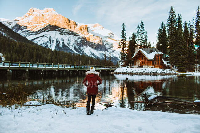 Back view of unrecognizable female tourist in outerwear holding hands in pockets and admiring wooden shack and mountain ridge while walking on snowy coast of Emerald Lake on winter day in British Columbia, Canada — Stock Photo