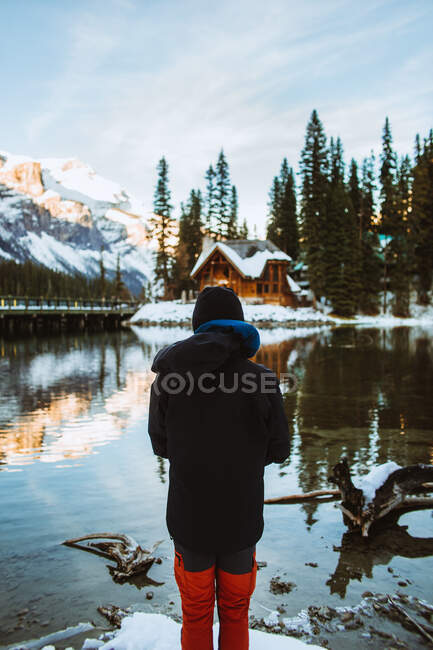 Rückansicht eines unkenntlich gemachten männlichen Touristen in Oberbekleidung, der an einem Wintertag in British Columbia, Kanada, an der schneebedeckten Küste des Emerald Lake steht und Holzhütten und Bergrücken bewundert — Stockfoto