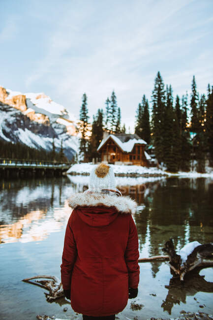 Back view of unrecognizable female tourist in outerwear admiring wooden shack and mountain ridge while standing on snowy coast of Emerald Lake on winter day in British Columbia, Canada — Stock Photo