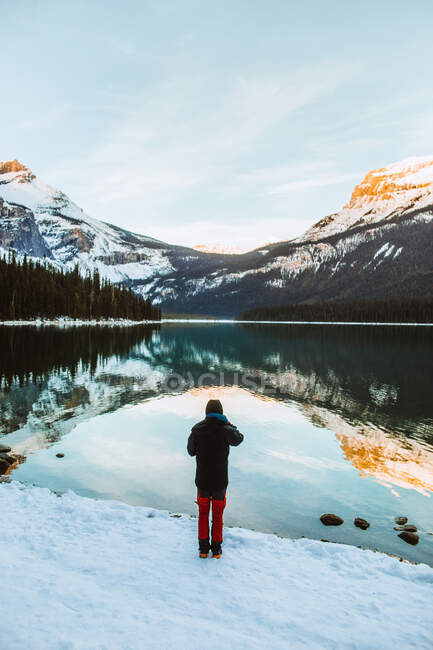 Vue arrière d'une personne anonyme en vêtements chauds debout sur du bois flotté près de l'eau calme du lac Emerald contre une randonnée en montagne enneigée et une forêt de conifères lors d'une journée d'hiver en Colombie-Britannique, Canada — Photo de stock