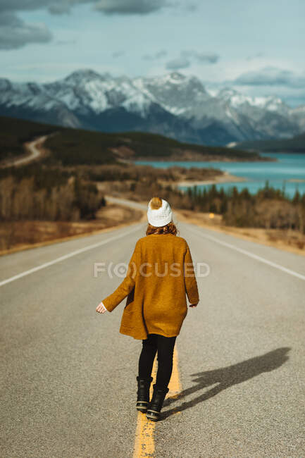 Back view of unrecognizable female tourist in casual clothes walking on David Thompson Highway near abraham lake against snowy mountains in Alberta, Canada — Stock Photo