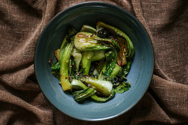 Top view of ceramic bow of yummy fried bok choy with sesame placed on brown fabric — Stock Photo