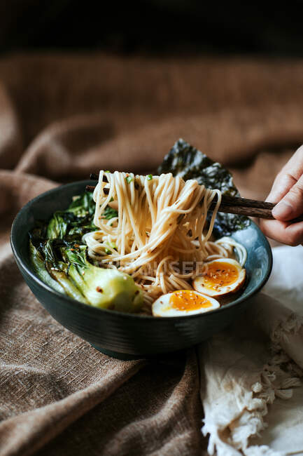 Unrecognizable person taking noodles with chopsticks while eating delicious ramen from bowl placed on brown fabric — Stock Photo