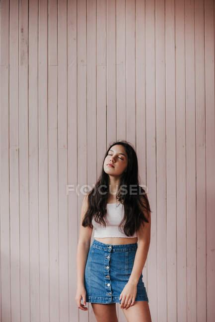 Calm dreamy female teenager with long dark hair wearing casual crop top and denim skirt standing with eyes closed against plank wall — Stock Photo