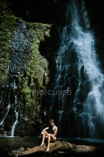 Молодая босая женщина-авантюристка в бикини сидит на камне против крутой скалы с зеленым мхом и плескающимся водопадом в солнечный день в муниципальном парке отдыха Лос-Чоррос в Коста-Рике — стоковое фото