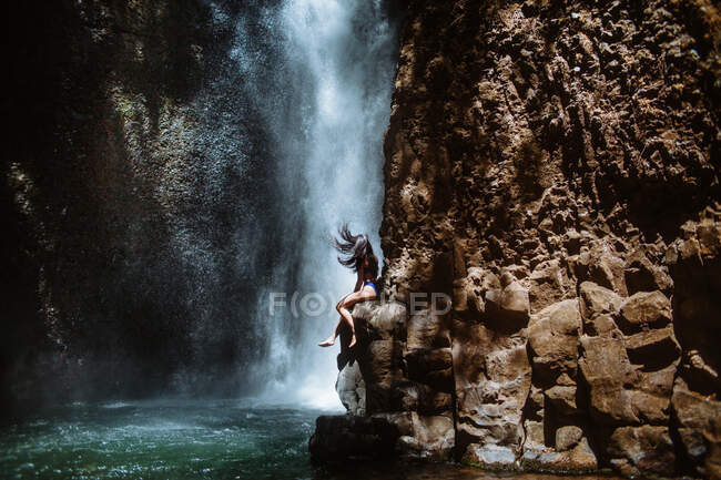 Вид сбоку на неузнаваемую женщину, расслабляющуюся и наслаждающуюся свежим воздухом, сидя на неровном скальном склоне у водопада Спилинг в солнечный день в муниципальном парке отдыха Лос-Анджелеса в Коста-Рике — стоковое фото