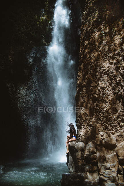 Side view of unrecognizable female traveler relaxing and enjoying fresh air while sitting on rough rocky slope near splashing waterfall in sunny day in Los Chorros Municipal Recreation Park in Costa Rica — Stock Photo