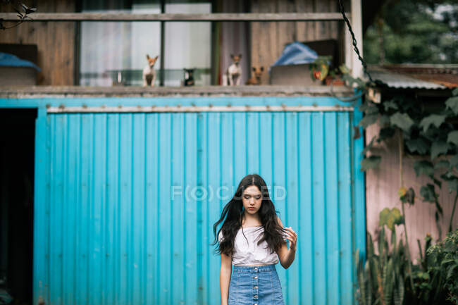 Sad pensive teen female in casual clothes touching hair while standing against countryside house with blue shutters and little dogs — Stock Photo