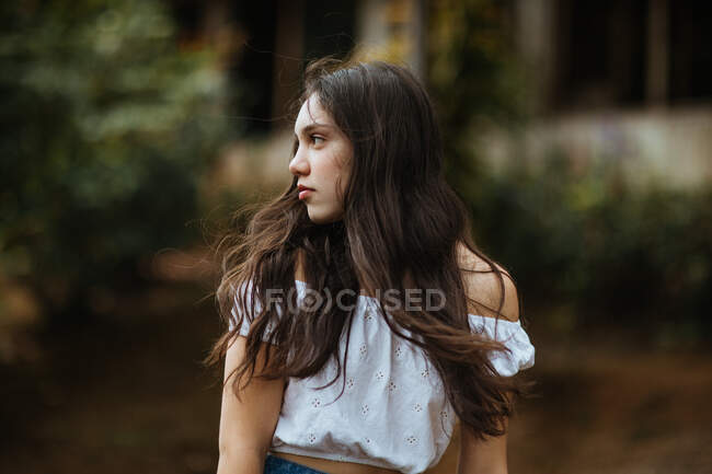 Young long haired brunette adolescent in white summer top looking away thoughtfully while standing against blurred greenery in nature — Stock Photo