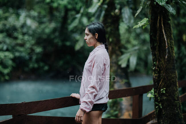 Vista lateral del joven explorador femenino reflexivo mirando hacia otro lado con interés mientras está parado en una pasarela de madera sobre el río Celeste con agua turquesa rodeada de exuberante vegetación tropical verde en Costa Rica - foto de stock