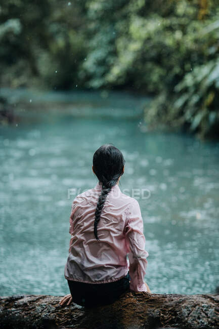Обратный вид на неузнаваемую женщину-путешественницу, сидящую на берегу спокойной реки Селеста с бирюзовой водой, текущей через зеленые тропические леса, расслабляясь и наслаждаясь уединением во время летнего путешествия в Коста-Рике — стоковое фото