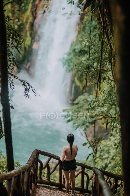 Desde arriba vista trasera de una turista irreconocible parada en el mirador y observando un paisaje pintoresco con salpicaduras de cascada y agua turquesa del río Celeste entre exuberante follaje verde en Costa Rica - foto de stock