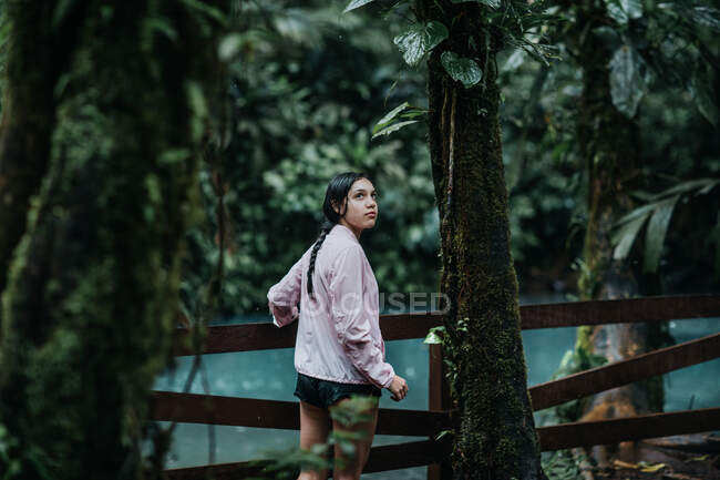 Vista posteriore di giovane esploratore femminile guardando oltre le spalle con interesse mentre in piedi su un ponte pedonale in legno sul fiume Celeste con acqua turchese circondata da una lussureggiante vegetazione tropicale verde in Costa Rica — Foto stock