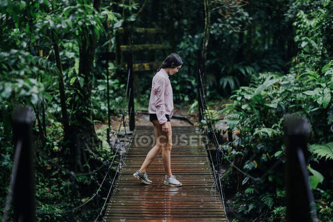 Ganzkörper-Seitenansicht einer jungen Reisenden, die während eines Sommerabenteuers in Costa Rica auf einer schmalen Hängebrücke über dem Fluss Celeste mit üppig grüner tropischer Vegetation steht — Stockfoto