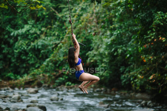 Vista lateral de una joven aventurera en traje de baño saltando sobre un arroyo que fluye a través de una exuberante selva tropical durante las vacaciones de verano en la provincia de Alajuela, Costa Rica - foto de stock