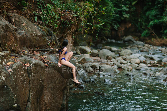 Full body side view of unrecognizable female traveler in bikini sitting on rocky shore of river flowing through green tropical forest during summer journey in Alajuela province of Costa Rica — Stock Photo