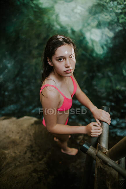 From above side view of young female traveler in pink bikini looking at camera while standing near railing on rocky shore of natural pool in Rincon de la Vieja National Park in Costa Rica — Stock Photo