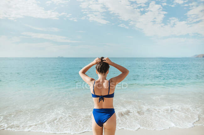 Обратный вид на неузнаваемую стройную брюнетку в голубых купальниках, трогающую длинные волосы, стоя в одиночестве на песчаном пляже против размахивающего океана и холмистого побережья во время летних каникул в Тамариндо в Коста-Рике — стоковое фото