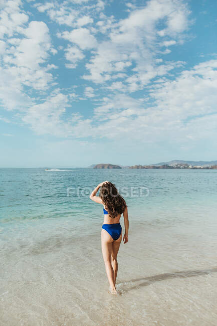 Rückansicht einer nicht wiederzuerkennenden schlanken Brünetten in blauer Badebekleidung, die lange Haare berührt, während sie allein am Sandstrand vor dem wogenden Meer und der hügeligen Küste während der Sommerferien in Tamarindo in Costa Rica steht — Stockfoto