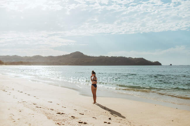 Corpo pieno di giovane donna in bikini in piedi da sola sulla spiaggia sabbiosa vuota vicino all'oceano ondulato mentre si gode la giornata di sole durante le vacanze in Tamarindo Costa Rica — Foto stock