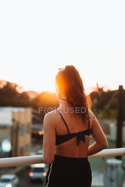 Vista trasera de una mujer irreconocible en traje de verano parada cerca de la barandilla y admirando la puesta de sol mientras descansa en la ciudad nocturna de Costa Rica - foto de stock