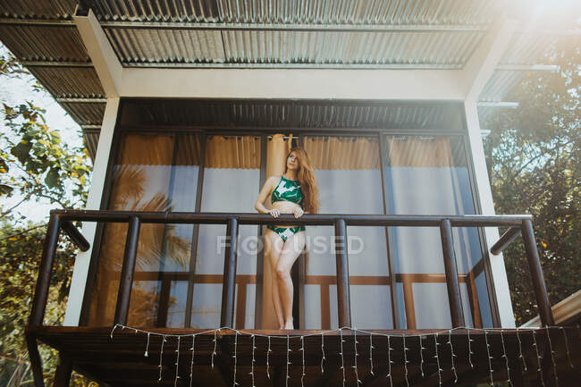 Desde abajo cuerpo completo de joven viajera de pelo largo en traje de baño con estilo de pie en el balcón de la casa de playa ubicada cerca de árboles verdes en el día de verano en la ciudad de Uvita en Costa Rica - foto de stock