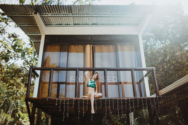 D'en bas corps complet de jeune voyageuse aux cheveux longs en maillots de bain élégants assis sur le balcon de la maison de plage située près des arbres verts dans la journée d'été dans la ville d'Uvita au Costa Rica — Photo de stock