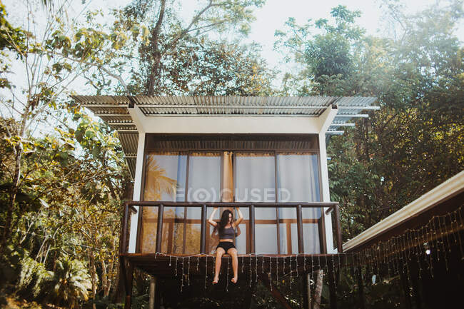 Нижче повного тіла молодих довговолосих подорожніх у стильному купальнику сидять на балконі пляжного будинку, розташованому біля зелених дерев влітку в місті Увіта, Коста-Рика. — стокове фото