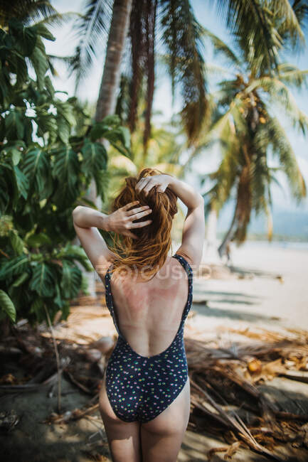 Vista posteriore di irriconoscibile rossa viaggiatore femminile in costume da bagno toccare i capelli mentre in piedi vicino a palme verdi sulla spiaggia di sabbia durante le vacanze estive in Costa Rica — Foto stock