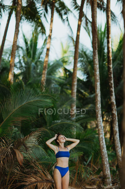 Mujer joven pacífica en traje de baño de pie con las manos en la cabeza y los ojos cerrados contra las palmas verdes altas durante las vacaciones de verano en la costa de Costa Rica - foto de stock