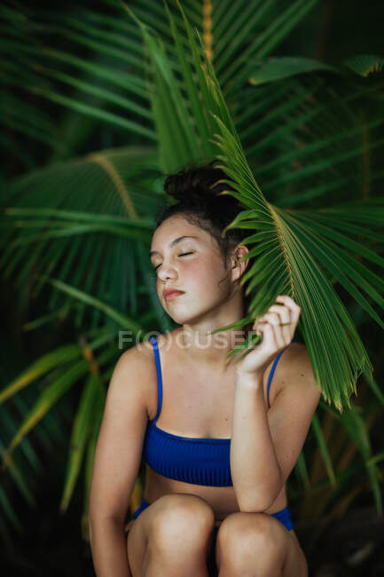 Charming young slim female in blue swimwear sitting with eyes closed on tree trunk under green palm foliage and looking at camera while enjoying summer vacation on beach of Costa Rica — Stock Photo