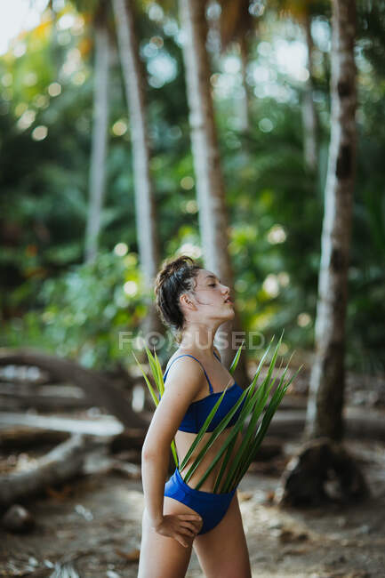 Jeune femelle en maillot de bain bleu posant le regard tourné vers l'extérieur avec des feuilles de palmier vert tout en se reposant sur une plage tropicale avec une végétation luxuriante pendant les vacances d'été au Costa Rica — Photo de stock