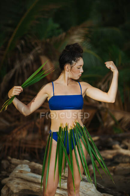 Femme en bikini et jupe en feuille de palmier montrant biceps debout dans un parc tropical à Uvita — Photo de stock