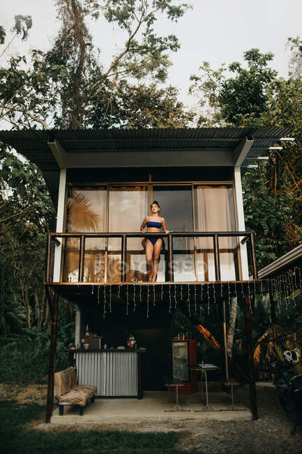 Desde abajo cuerpo completo de joven viajera de pelo largo en traje de baño con estilo de pie en el balcón de la casa de playa ubicada cerca de árboles verdes en el día de verano en la ciudad de Uvita en Costa Rica - foto de stock