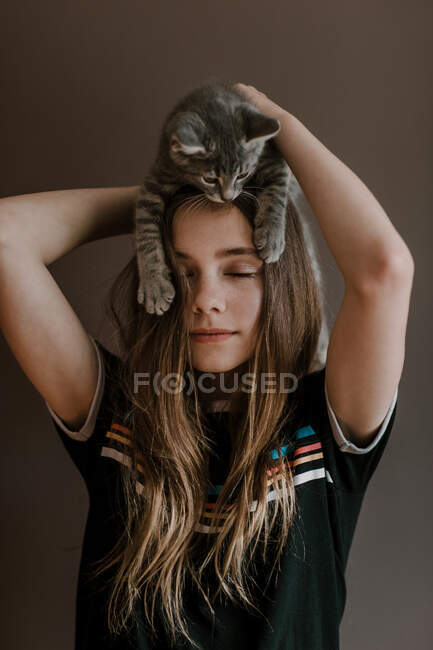 Мечтательная девочка-подросток с пушистым милым котом на голове на коричневом фоне в студии — стоковое фото