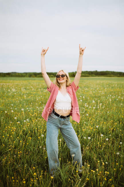 Joyeux jeune femme debout dans la prairie en été et montrant signe de roche tout en regardant la caméra — Photo de stock