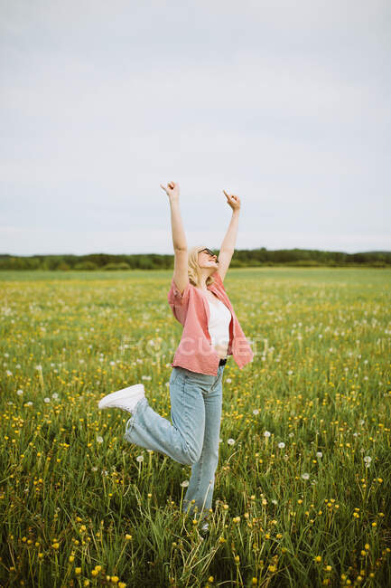 Joyeux jeune femme debout dans la prairie en été et montrant signe de roche tout en regardant la caméra — Photo de stock