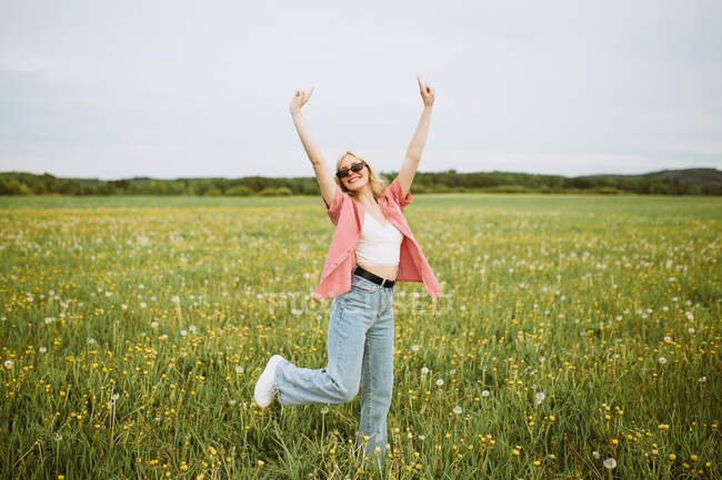 Feliz joven hembra de pie en el prado en verano y mostrando el signo de roca mientras mira la cámara - foto de stock