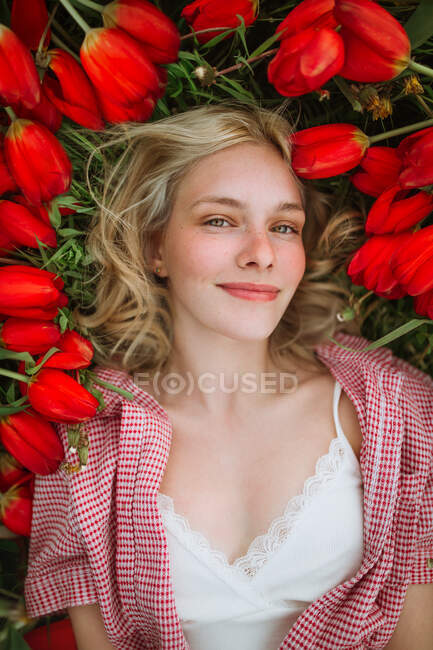 Vue du dessus de la femelle positive couchée dans le champ avec des fleurs de tulipes rouges et regardant la caméra — Photo de stock