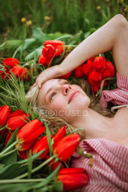Vista superior de hembra positiva acostada en el campo con flores de tulipán rojo y mirando hacia otro lado - foto de stock