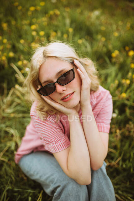 Молодая безмятежная женщина в модном наряде летом сидит на цветущем лугу и смотрит в камеру — стоковое фото