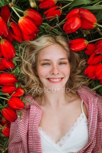 Visão superior da fêmea positiva deitada em campo com flores de tulipa vermelha e olhando para a câmera — Fotografia de Stock
