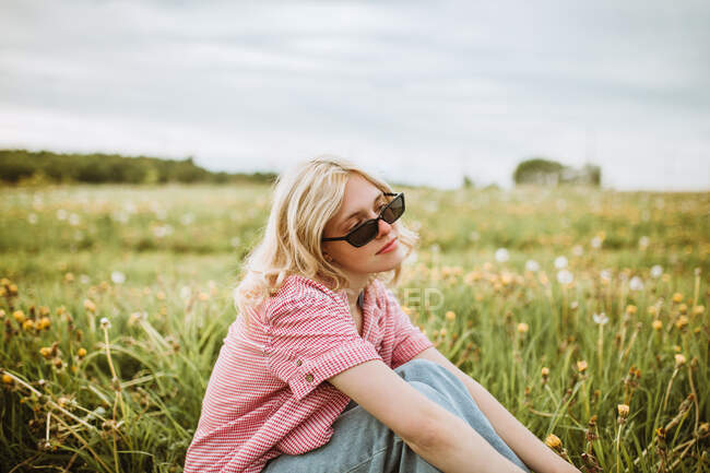 Vista lateral de la joven hembra serena en traje de moda sentada en el prado floreciente en verano y mirando hacia otro lado - foto de stock