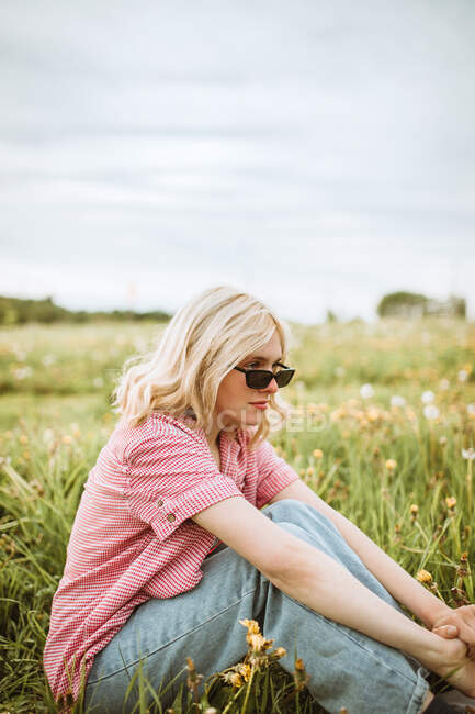 Vista lateral da jovem fêmea serena na roupa da moda sentada no prado florescente no verão e olhando para longe — Fotografia de Stock