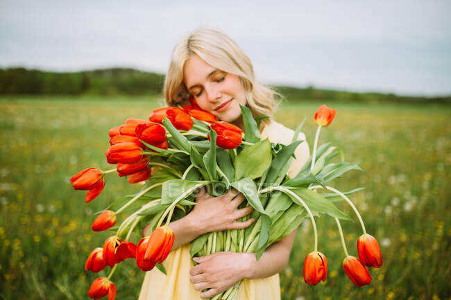 Conteúdo fêmea de vestido em pé com um monte de flores de tulipa vermelha no prado no verão com olhos fechados — Fotografia de Stock