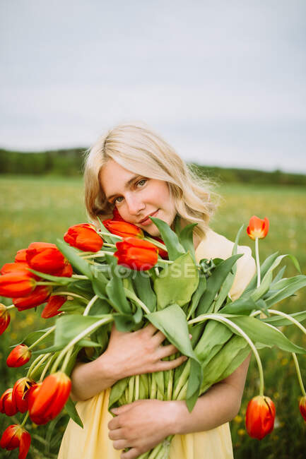 Zufriedene Frau im Kleid steht im Sommer mit einem Strauß roter Tulpenblumen auf der Wiese — Stockfoto