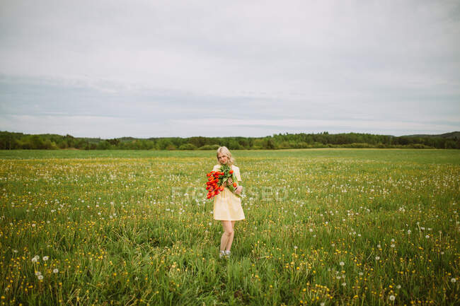 Contenido femenino en vestido de pie con ramo de flores de tulipán rojo en el prado en verano - foto de stock